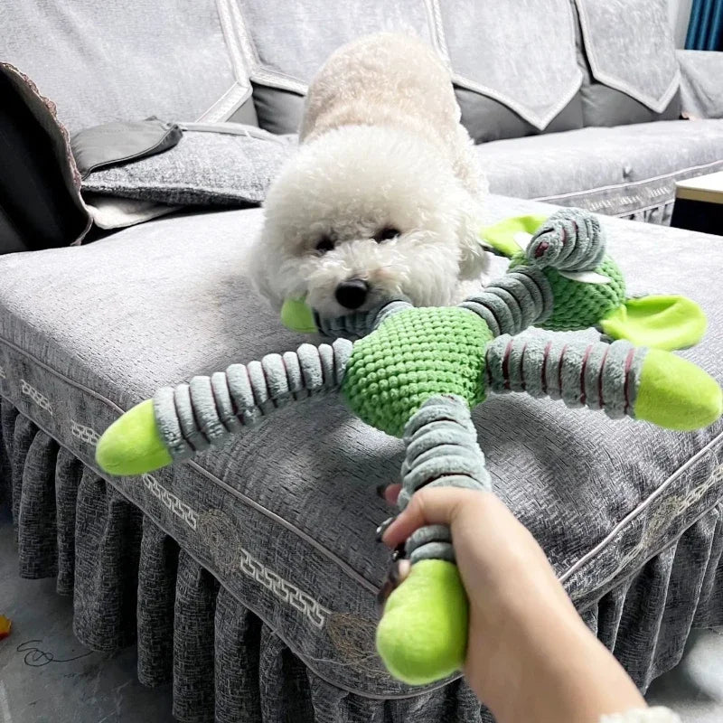 Ein weißer flauschiger Hundespielzeug mit grünen Akzenten wird von einer Person auf einem grauen Sofa gehalten.