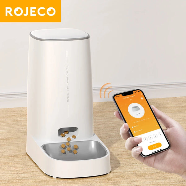 A white Rojeco smart pet feeder device with a feeding tray is shown next to a hand holding a smartphone.