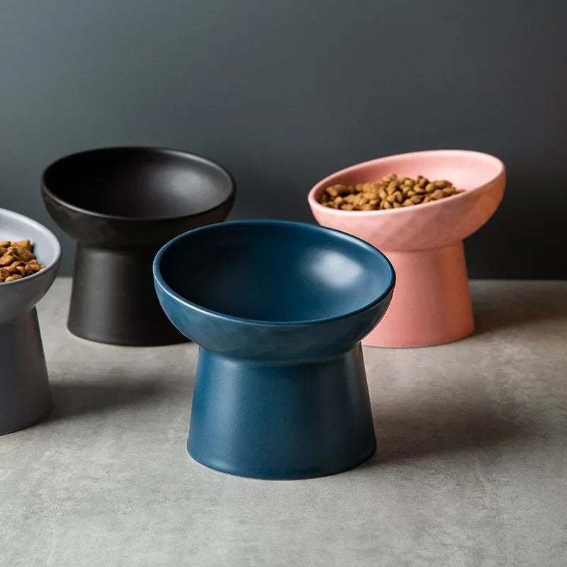 Three elevated ceramic food bowls in black, pink, and blue sit on a gray surface.