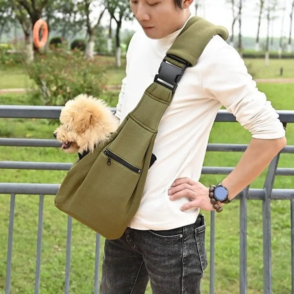 A person wearing a white shirt holds a olive green pet carrier bag with a safety harness and a fluffy dog.