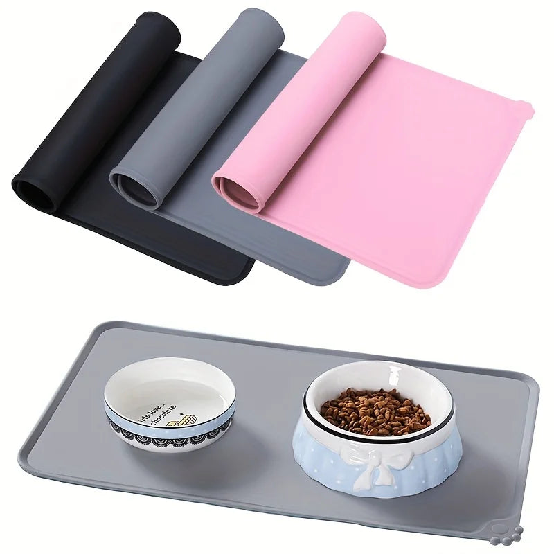 A pink silicone pet feeding mat is shown rolled up and a gray silicone feeding tray with a bowl of kibble.