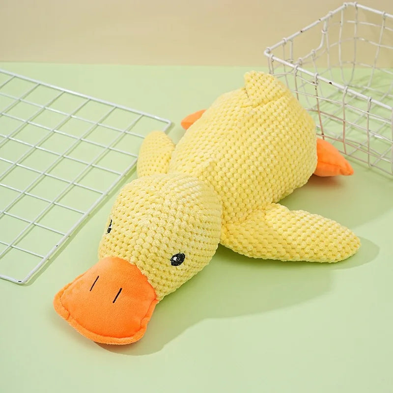 A yellow knitted plush toy shaped like a duck with orange feet resting on a green surface next to a wire mesh basket.
