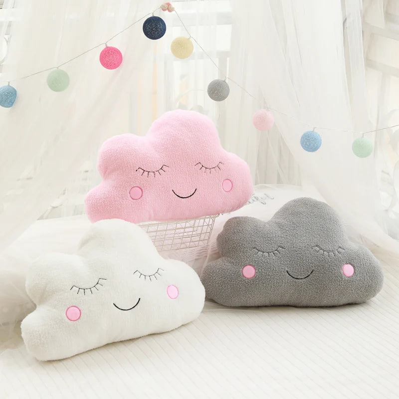 A collection of pink and gray cloud-shaped plush pillows with embroidered features, hanging from a string in a bright setting