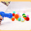 A hand holds a blue toy gun next to a pile of colorful, soft plush balls.
