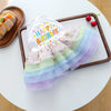 A white dog birthday suit with colorful tulle layers is displayed on a wooden tray next to a pastry.