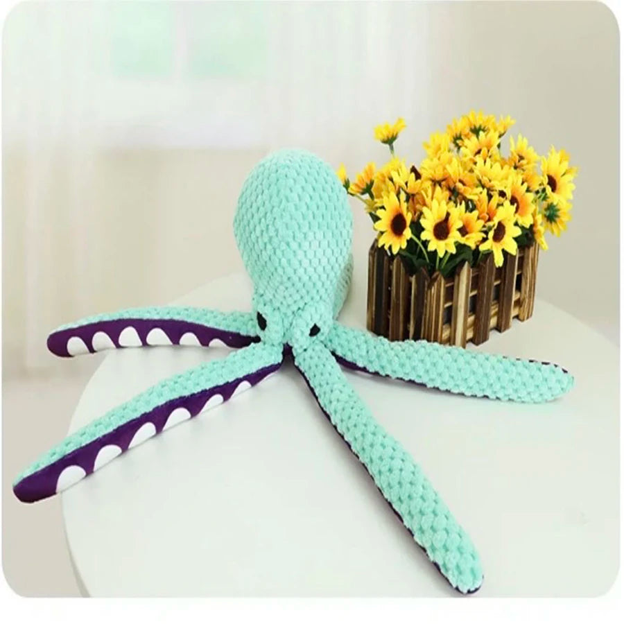 A handmade plush octopus toy with light blue body and purple accents lying next to a vase of yellow flowers.
