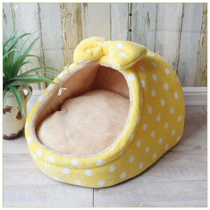 A yellow, polka-dotted cozy cave pet bed with a large bow, resting on a light wooden floor.