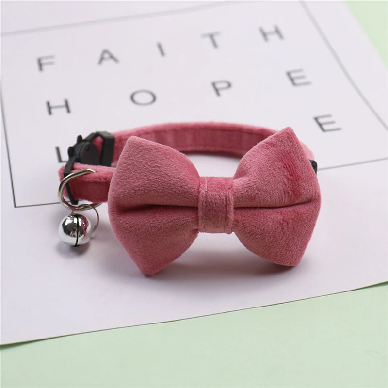 A pink plush velvet pet collar featuring a bow and a silver bell, resting on a white paper with faint text.