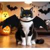 A black cat wearing a mask and wings sits on a wooden floor in front of a Halloween-themed background.