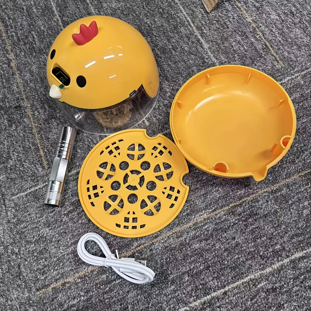 A yellow home egg incubator with a clear dome, an orange dish, and a yellow perforated tray, along with a white cord.