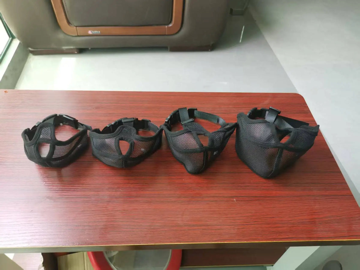 Four black adjustable breathable mesh dog muzzles laid out on a wooden table.
