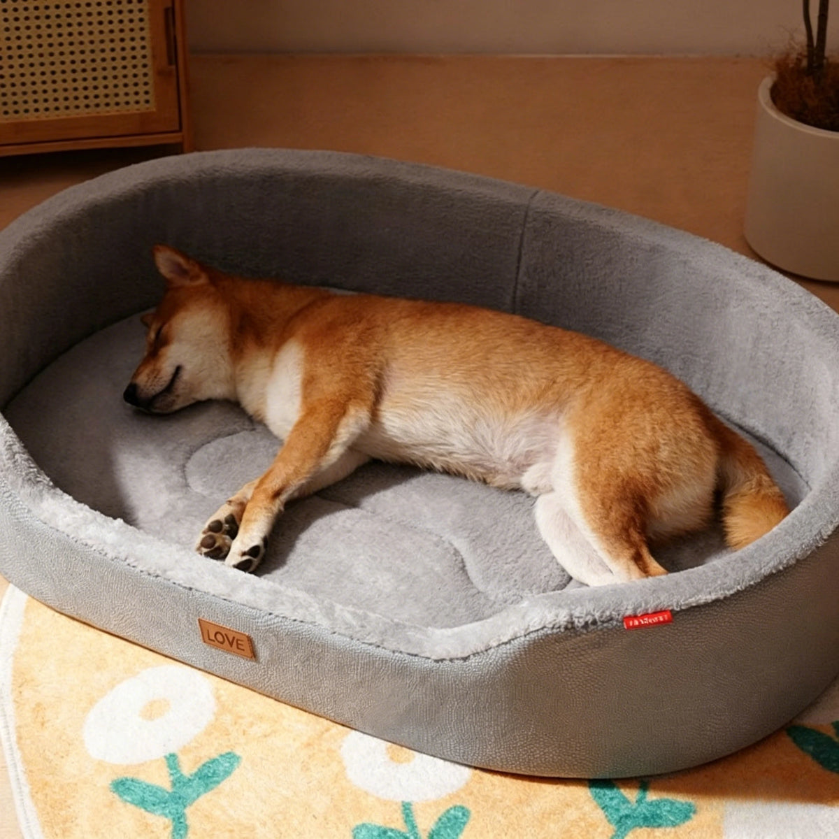 A light brown dog is resting inside a gray pet bed with a removable cushion on a yellow floral mat.