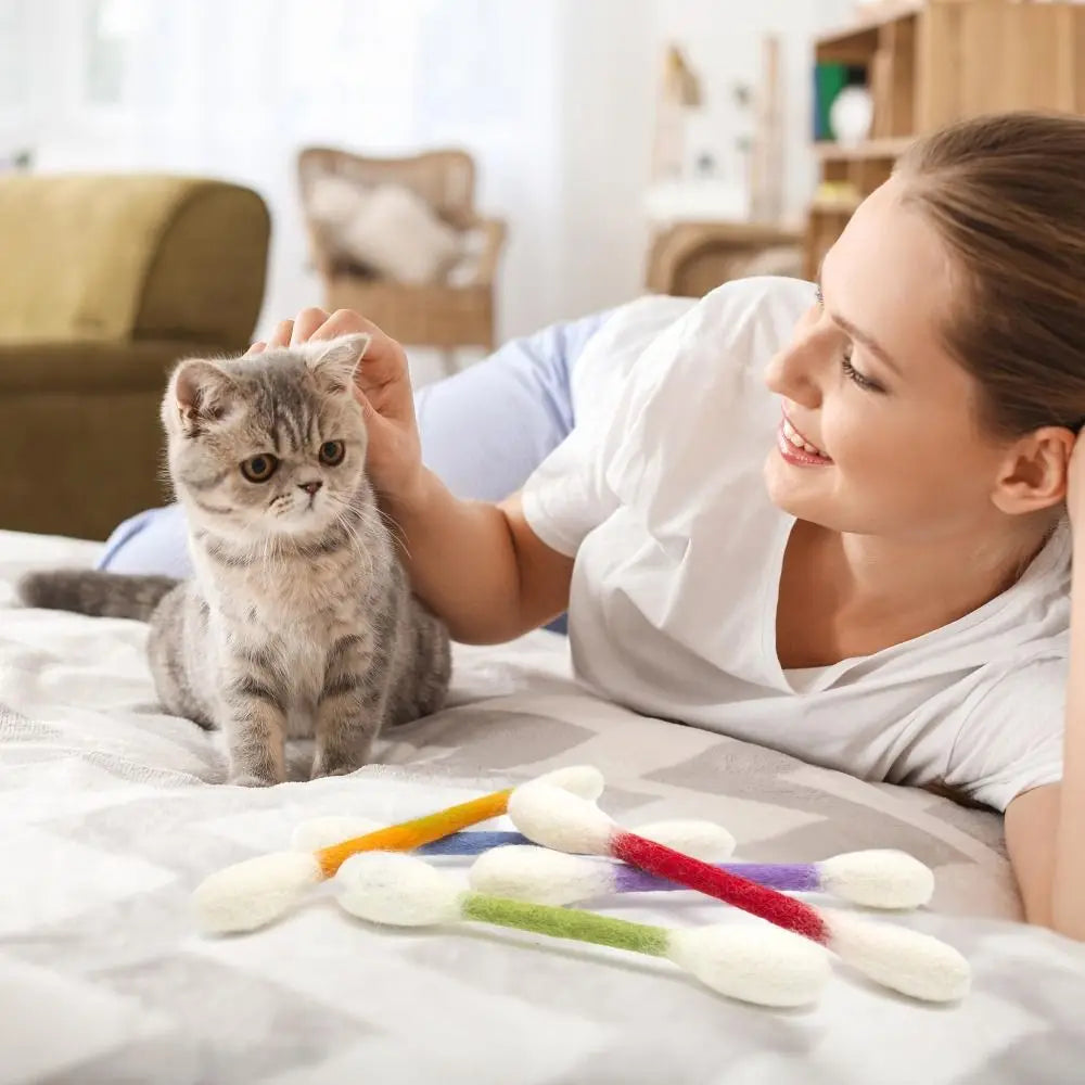 A woman gently pets a small tabby kitten while it plays with a pack of felt cotton swab toys.