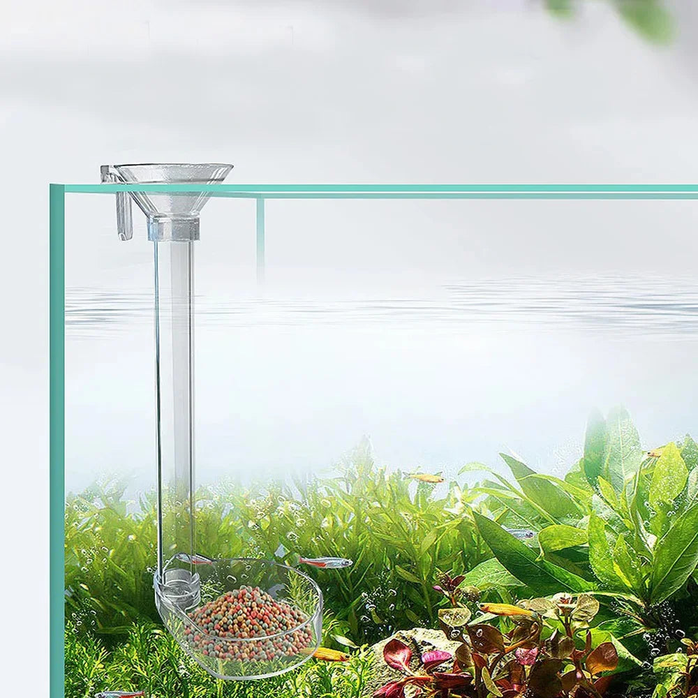 An adjustable aquarium feeder with a transparent tube is shown in a glass container surrounded by aquatic plants.