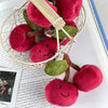 A pair of adorable cherry plush keychains in pink and green are displayed on a white mesh wire.