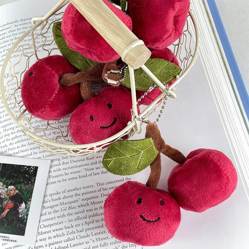 A pair of adorable cherry plush keychains in pink and green are displayed on a white mesh wire.