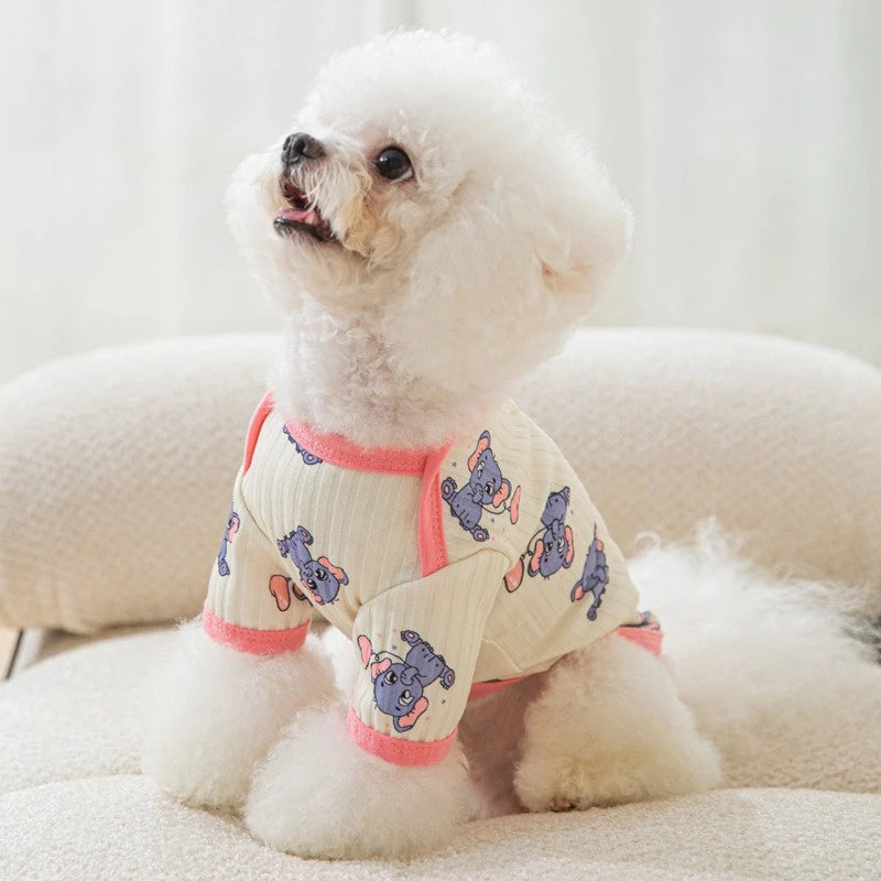 A fluffy white dog wearing a light-colored vest with a cartoon print of purple dogs.