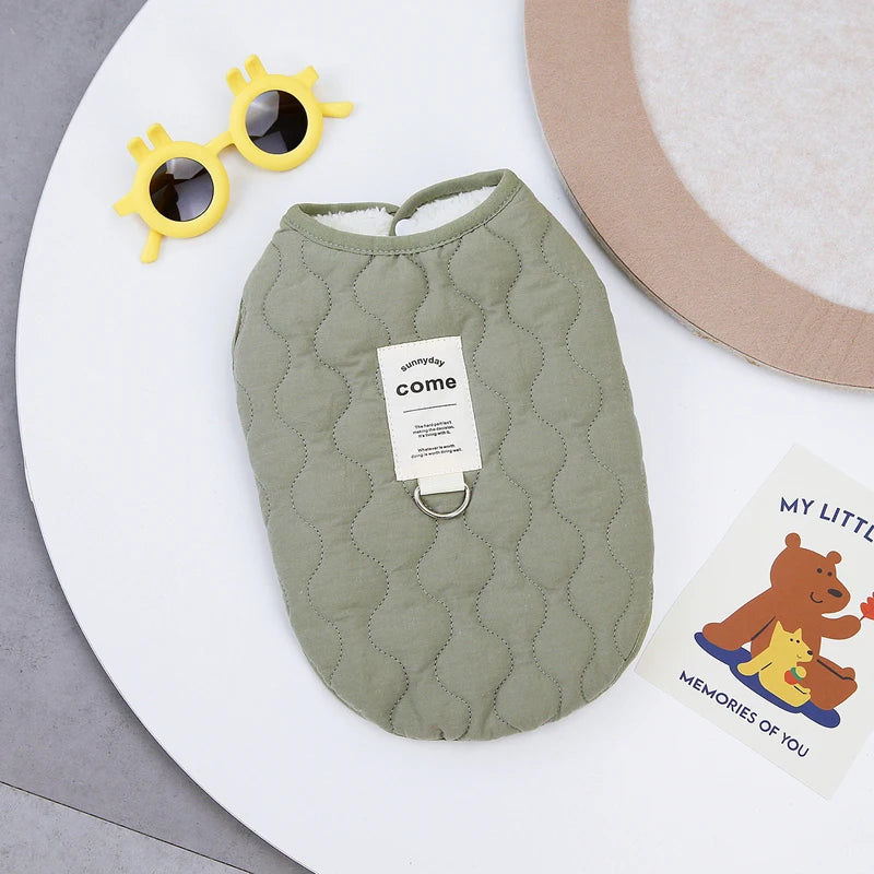 A soft sage green quilted pet vest with a white tag and a pair of yellow sunglasses is displayed on a white surface.