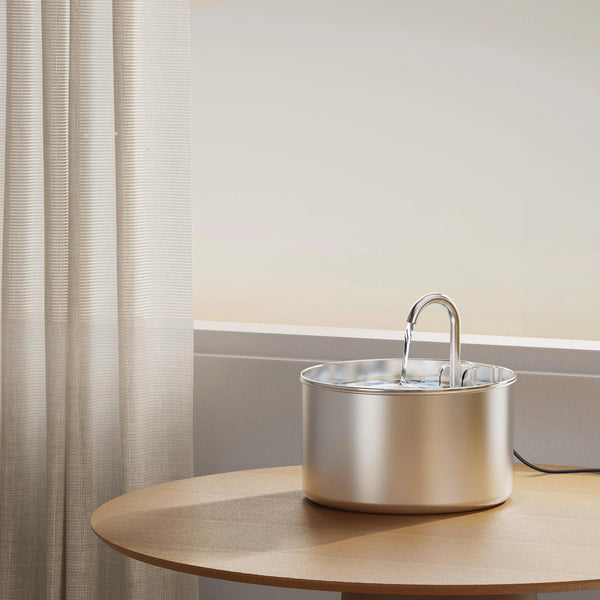 A stainless steel pet water fountain with a filter sits on a wooden table next to a sheer curtain.