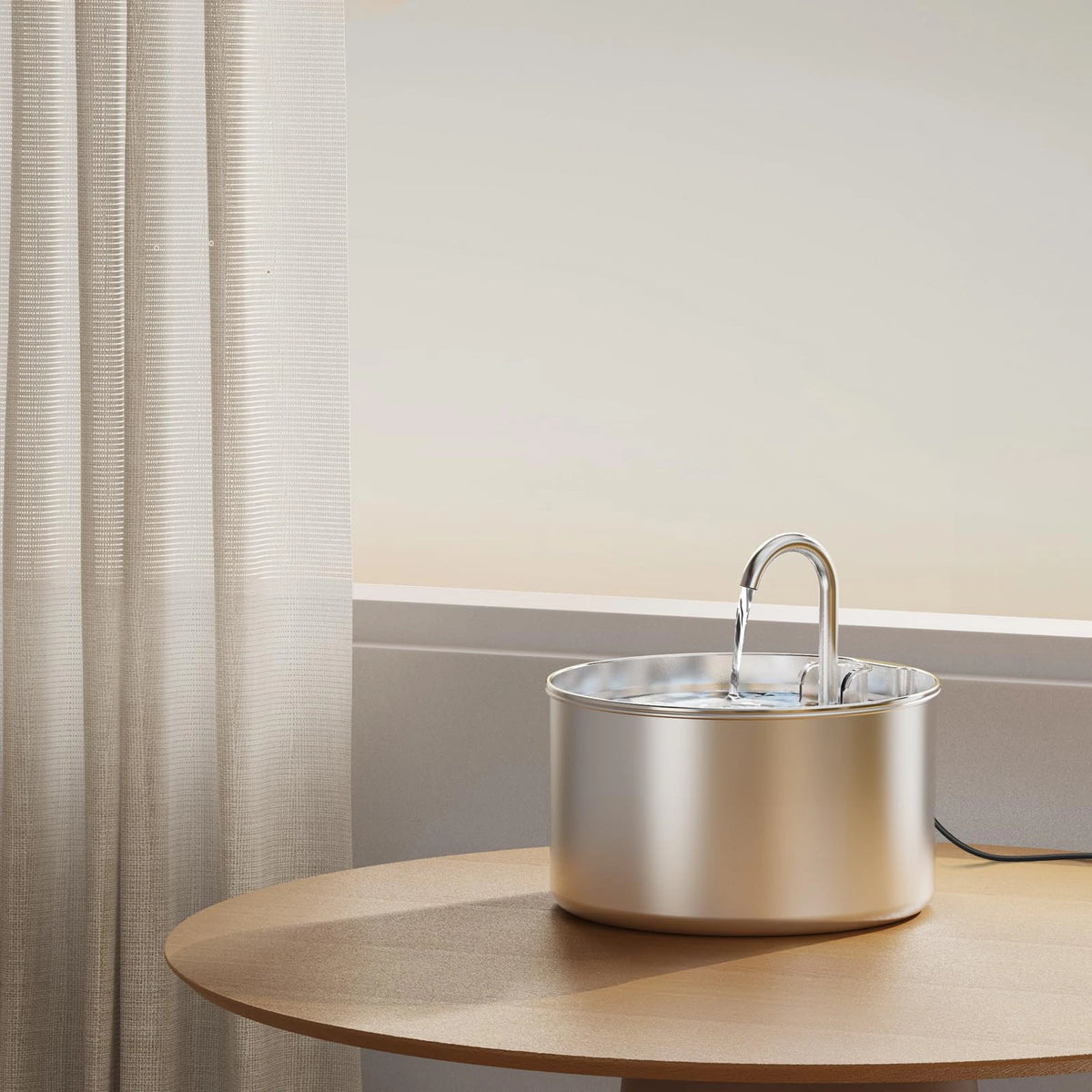 A stainless steel pet water fountain with a visible filter is placed on a wooden table next to a sheer curtain.