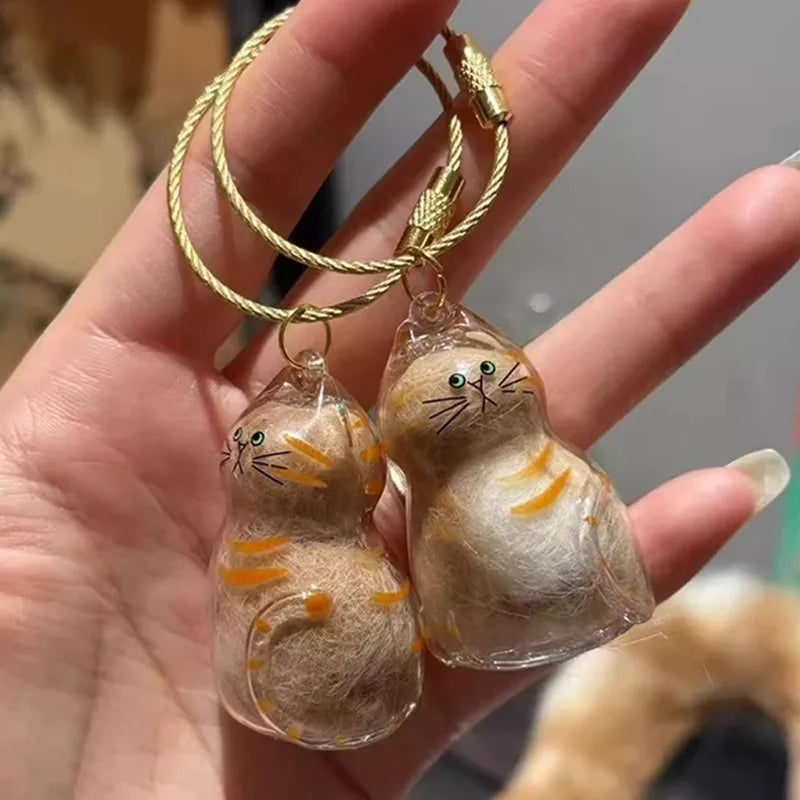 A small, round cat fur charm keychain with orange stripes is held in a hand.