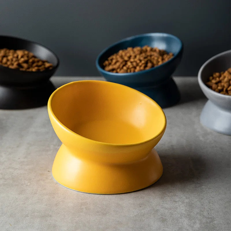 A bright yellow ceramic cat bowl with a raised stand is centered on a gray surface, with other bowls in the background.