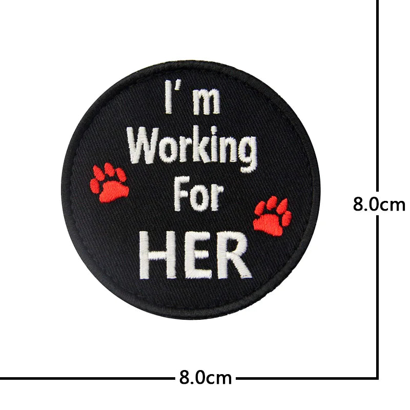 Black embroidered patch with white text I'm Working For HER and a red paw print.