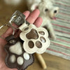 A leather paw print airtag keepsake with a cutout design is held in a hand next to a fluffy white dog.