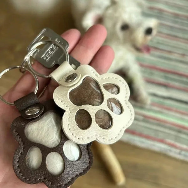 A leather paw print airtag keepsake with a cutout design is held in a hand next to a fluffy white dog.