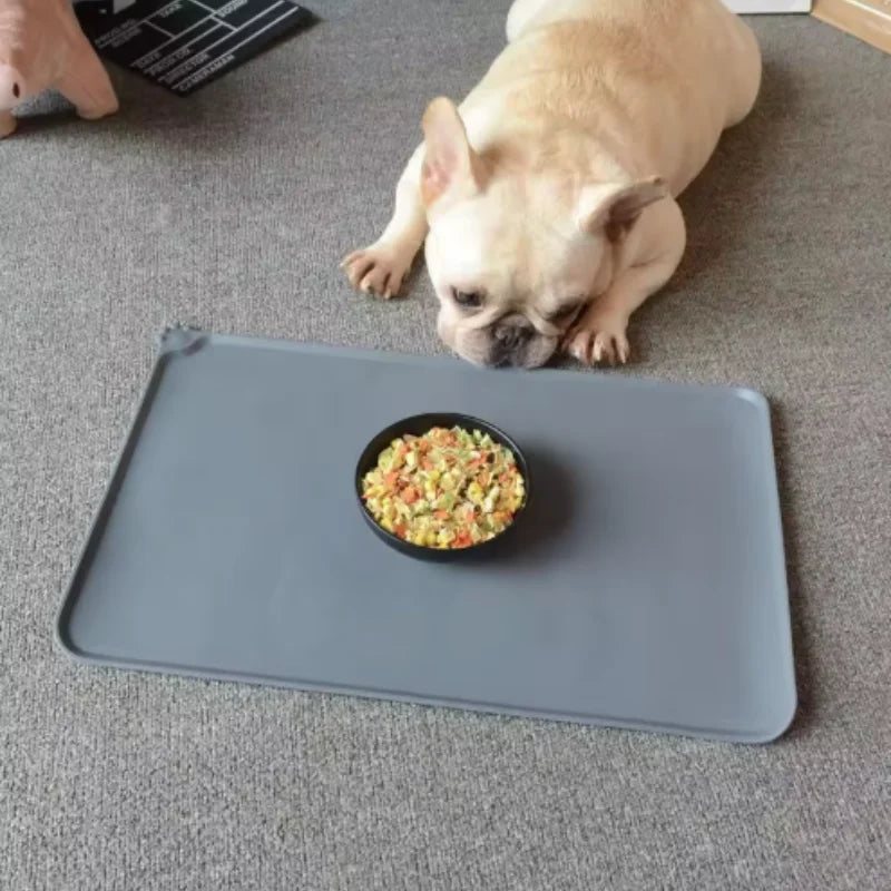 A light brown French Bulldog lies on a gray silicone pet feeding mat with a bowl of food.