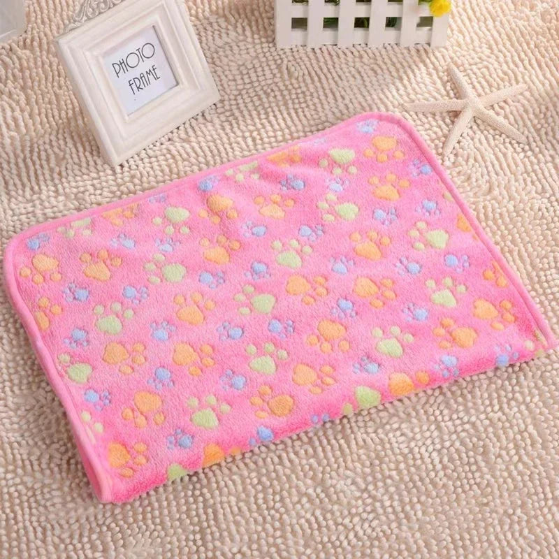 A pink flannel fleece pet blanket with a yellow and blue paw print design resting on a beige textured surface.