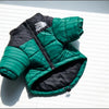 A green and black quilted coat for a dog is displayed against a white slatted background.