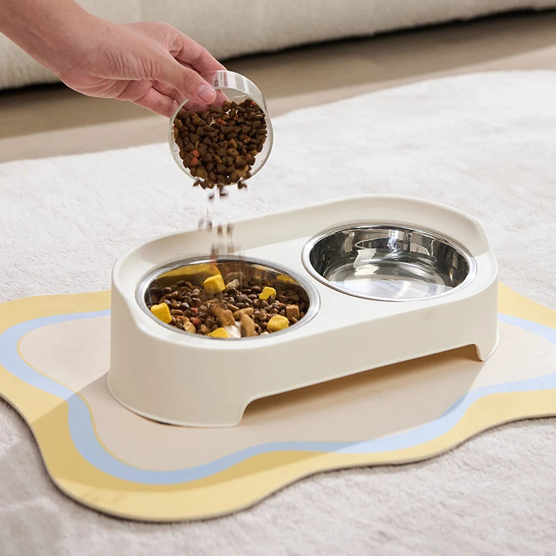 A hand is pouring dry pet food into a white feeder stand with two stainless steel bowls on a light-colored rug.