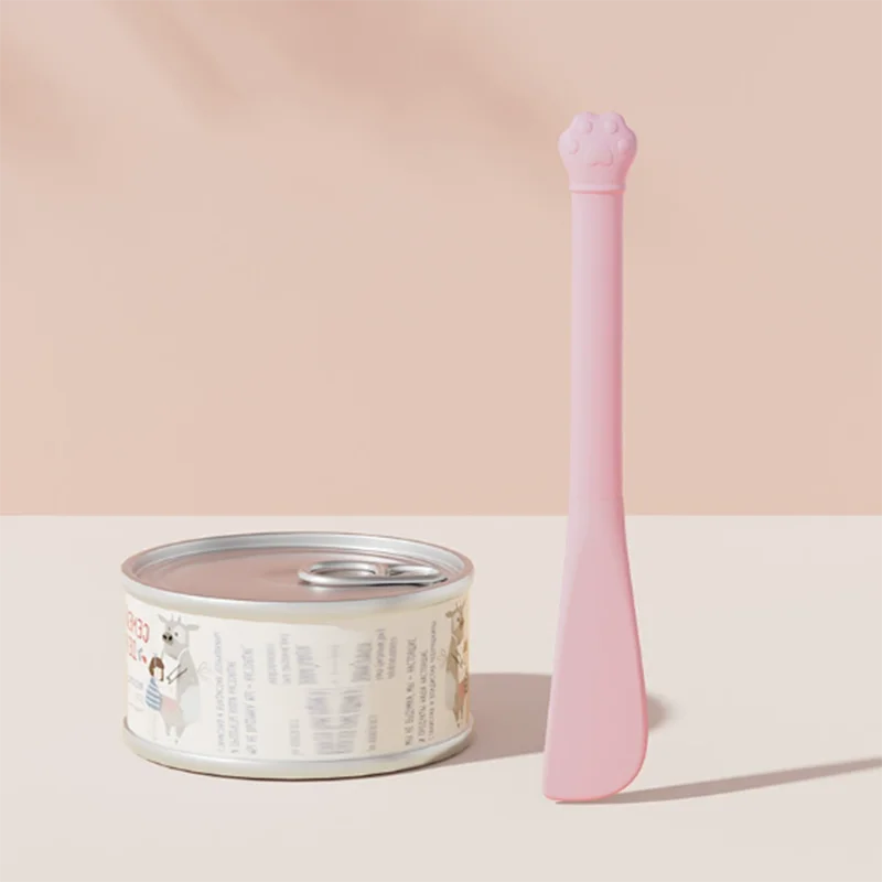 A pink multi-purpose pet feeding spoon with a built-in can opener and seal cap resting on a light surface.