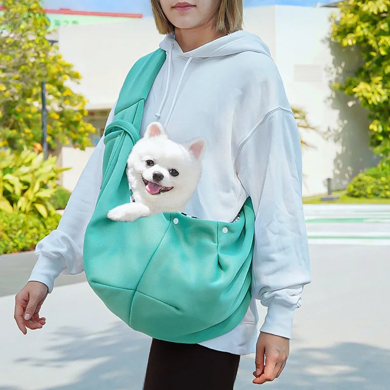 A person wearing a light blue hoodie is carrying a small white dog in a teal hands-free pet sling carrier.