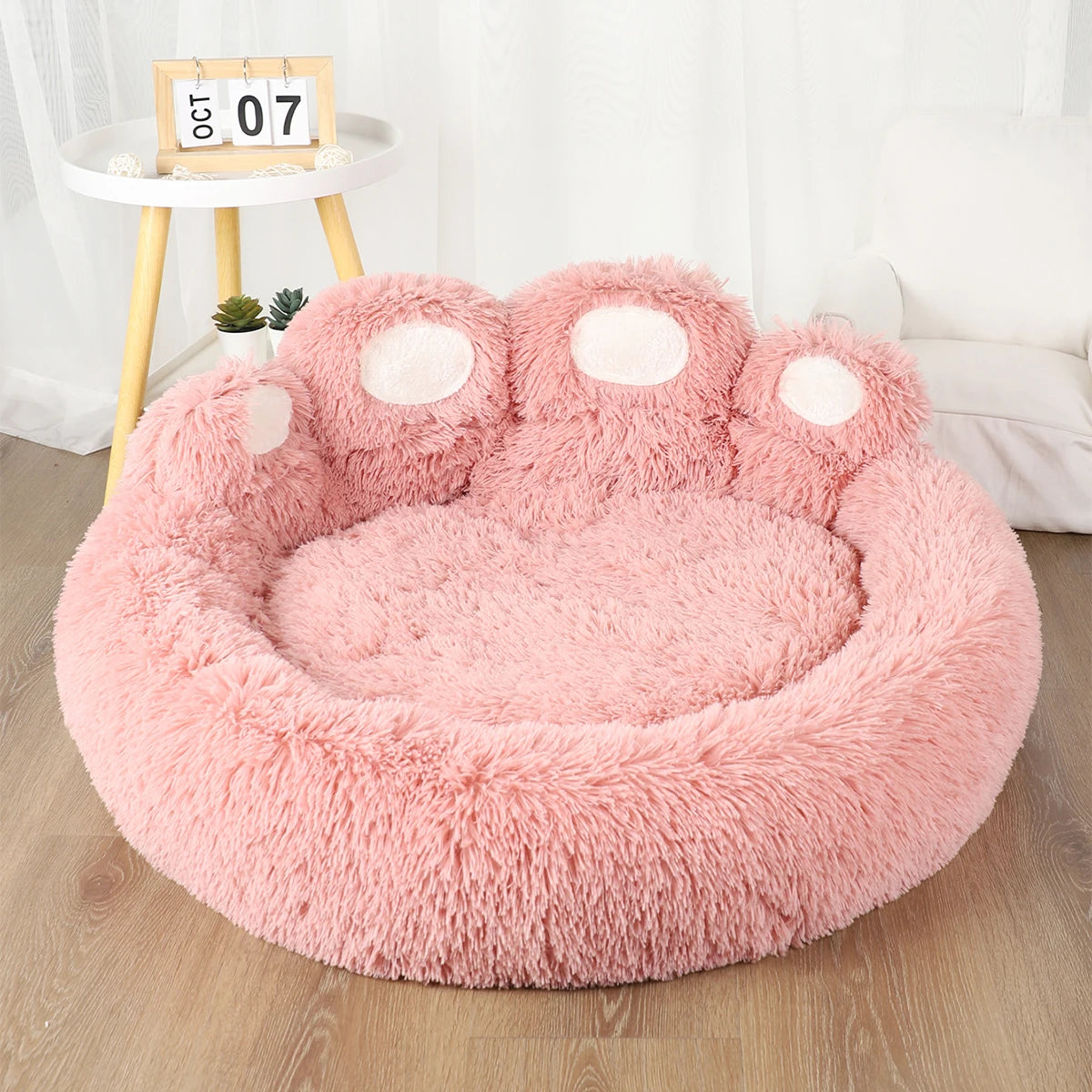 A pink plush fluffy dog bed shaped like a paw print resting on a wooden surface.