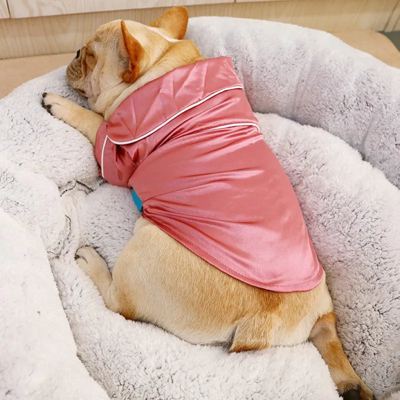 A light brown dog wearing a pink silk pajama top, relaxing on a fluffy white pet bed.