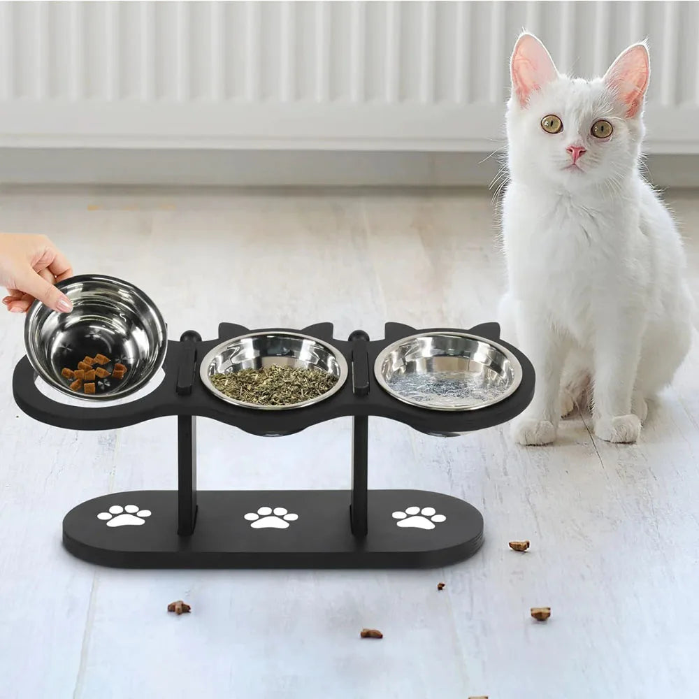 A black elevated cat food bowl stand holding three stainless steel bowls next to a white cat.