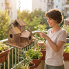 A young woman points at a rustic wooden bird house hanging on a balcony filled with potted plants.