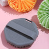 A gray silicone exfoliating brush is shown next to an orange and green exfoliating brush on a pink background.