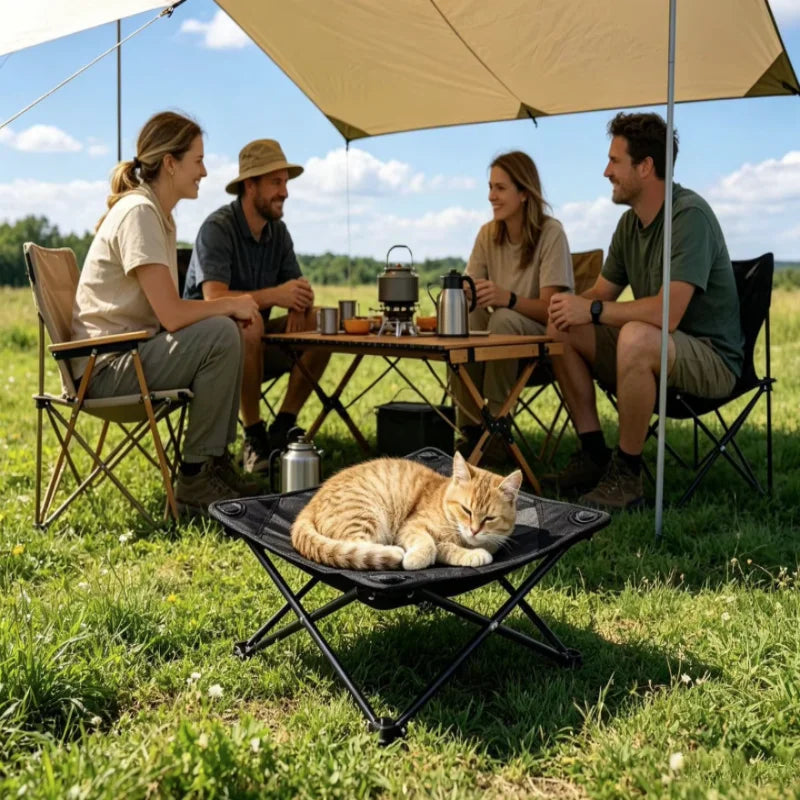 A group of people relaxing outdoors under a large umbrella with a cat resting on a black folding pet cot.