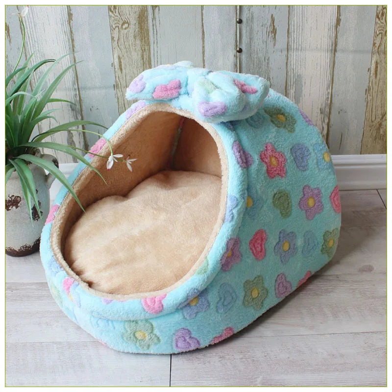 A light blue floral pet bed with a soft interior, resting on a wooden floor next to a potted plant.