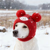 A white dog wearing a red knitted winter hat with bear ears in a snowy outdoor setting.