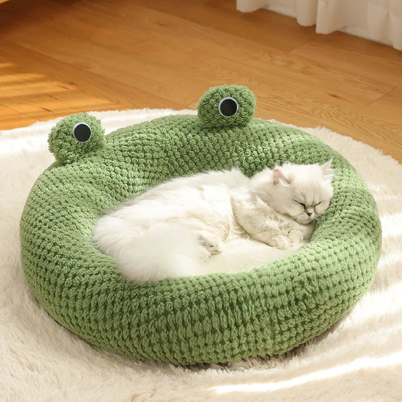 Un nido de peluche tejido verde con grandes ojos, sosteniendo un gatito blanco en una superficie suave y blanca.