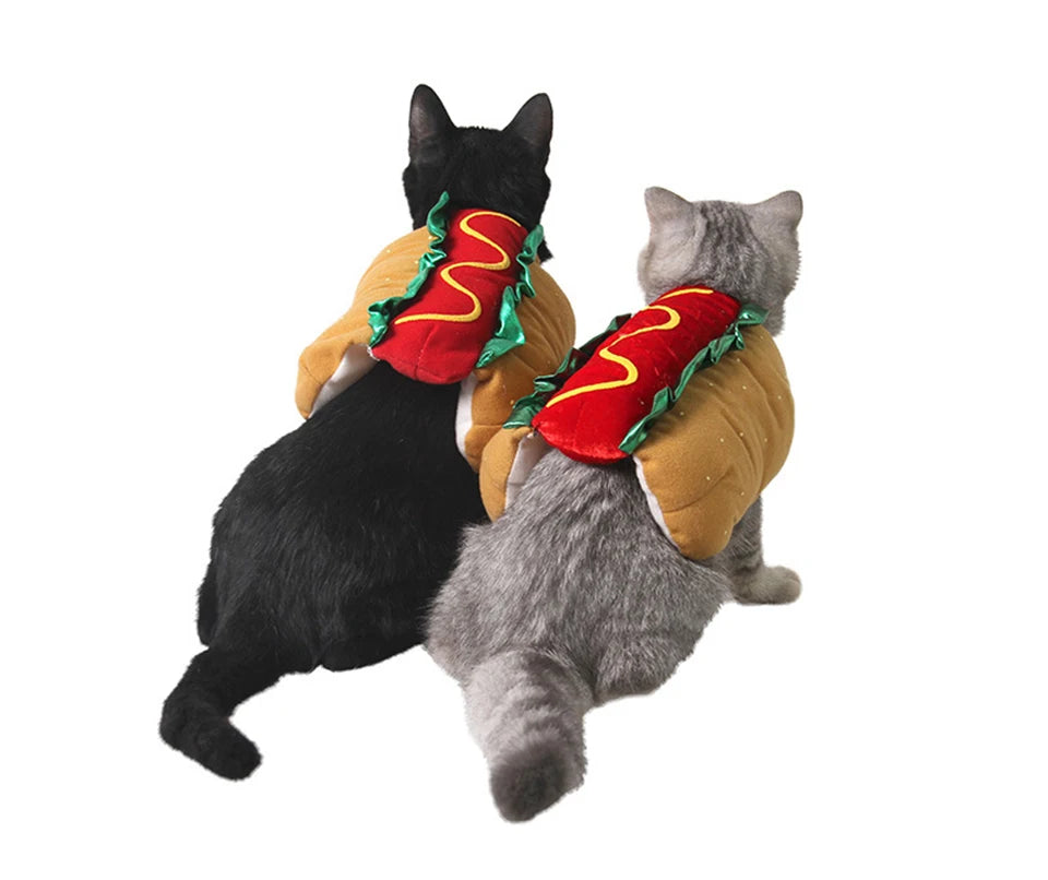 A black cat and a gray cat wearing cozy hot dog costumes with red and green toppings.