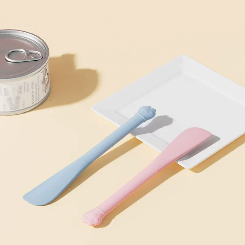 A light blue and pink multi-purpose pet feeding spoon with a built-in can opener resting on a white plate.