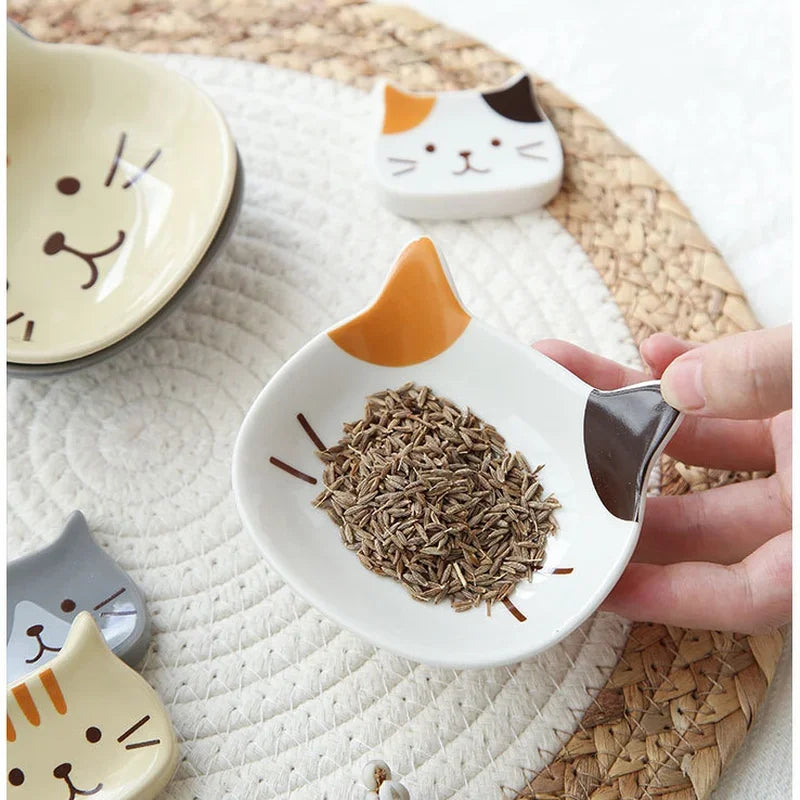 A hand holds a white ceramic cat-shaped sauce dish filled with dried herbs on a woven mat.