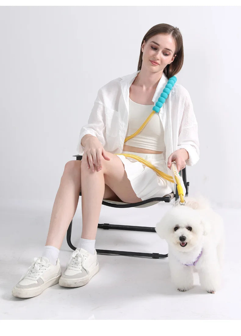 A woman is sitting on a chair with a white shirt and shorts, holding an adjustable hands-free dog leash.