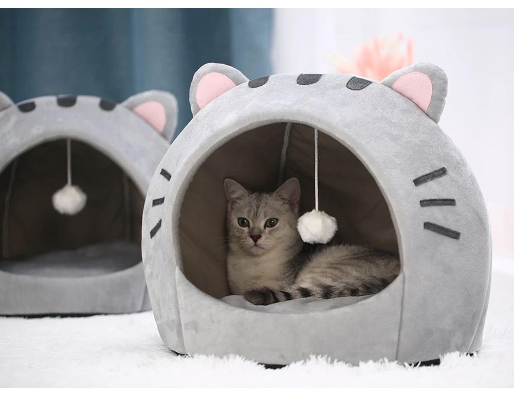Un lit grotte gris pour chat avec ouvertures d'oreilles et un chaton duveteux qui dort à l'intérieur.