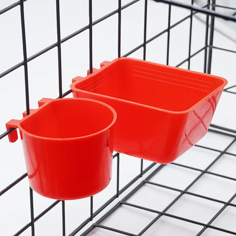 A bright red plastic bird feeder and water bowl are shown hanging in a black wire mesh cage.